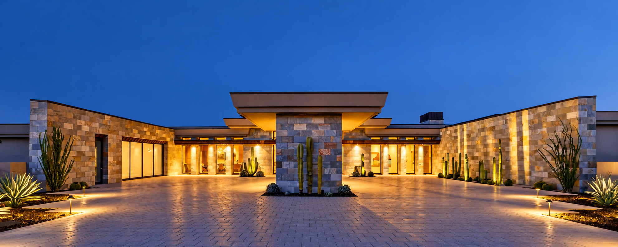 Collina, a modern single-story desert home in Scottsdale, Arizona, at blue hour, with a stone-clad entry pavilion, glowing windows, lit desert landscaping and a paver motor court